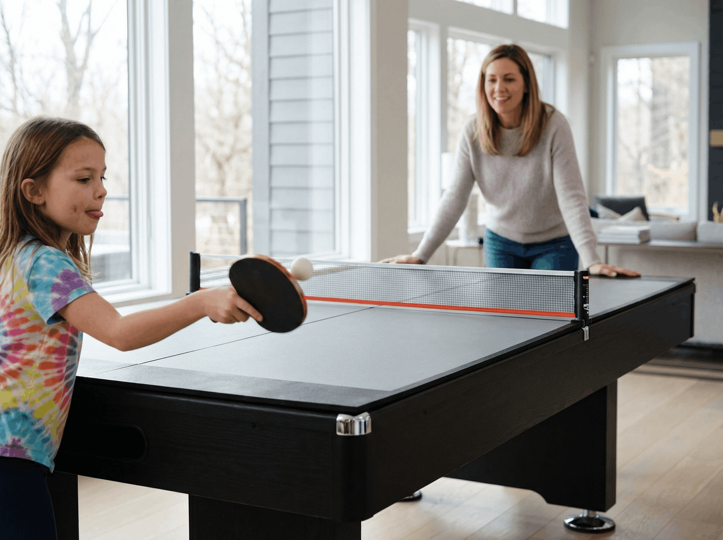 Two people playing ping pong in a bright room with large windows.